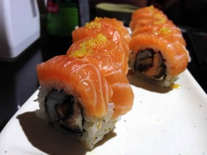 Skin Sushic: Uramaki de pele de salmão grelhada, com uma fatia de salmão em volta, raspas de limão e molho de maracujá.